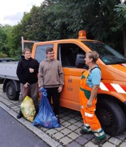 Zwei Jugendliche mit vollen Müllsäcken und eine Mitarbeiterin des Bauhofs in orangefarbener Arbeitskleidung stehen vor einem Fahrzeug der Stadt Meißen.