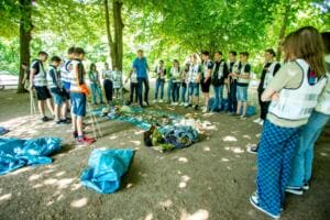 Viele Schülerinnen und Schüler in Warnwesten stehen im Kreis um ausgebreitete Planen mit dem gesammelten Müll, in der Mitte erklärt ein Erwachsener.