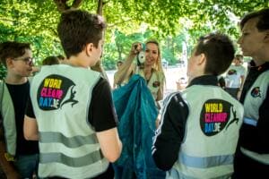 Eine Betreuerin verteilt Müllsäcke an eine Gruppe Schüler in World-Cleanup-Day-Warnwesten im Schatten von Bäumen.