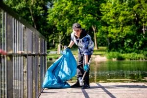 Ein lächelnder Junge mit Kappe hebt auf einem Holzsteg am Teich einen Plastikbecher mit der Greifzange auf und hält einen blauen Müllsack.
