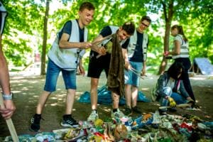 Jugendliche in Warnwesten sortieren lachend mit Greifzangen einen großen Haufen gesammelten Mülls auf einer Plane, darunter ein alter Lappen.