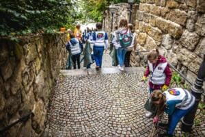 Mehrere Schülerinnen und Schüler in Warnwesten sammeln auf einer gepflasterten Treppe zwischen alten Natursteinmauern Müll.