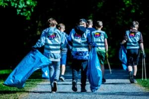 Fünf Schüler in World-Cleanup-Day-Warnwesten ziehen von hinten mit blauen Müllsäcken und Greifzangen einen Parkweg entlang.