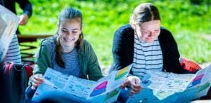Zwei lächelnde Schülerinnen sitzen im Grünen und lesen einen großen bunten Faltplan zum Thema Müll.
