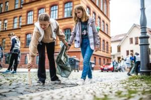 Zwei Schülerinnen in Warnwesten suchen mit Greifzange auf einer Kopfsteinpflasterstraße vor einem Backstein-Schulgebäude nach Müll.
