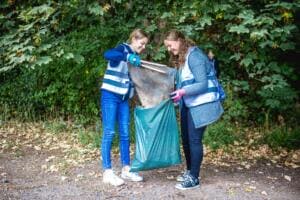Zwei Schülerinnen in Warnwesten füllen lachend mit Greifzangen einen großen Fund in einen grünen Müllsack, im Hintergrund Bäume.