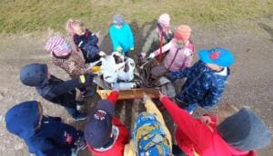 Von oben fotografiert stehen Kindergartenkinder in bunten Jacken im Kreis um einen Bollerwagen mit gesammeltem Müll und zeigen darauf.