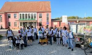 Eine große Gruppe von Erwachsenen und Kindern in weißen Warnwesten steht mit Greifzangen, Müllsäcken und einem Bollerwagen vor dem rosafarbenen Bahnhofsgebäude von Mahlow.