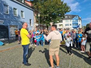 Zwei Männer stehen sich auf einer gepflasterten Straße gegenüber und sprechen, dahinter wartet eine große Gruppe aus Kindern und Erwachsenen mit Greifzangen und Warnwesten.