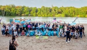 Weit über hundert Menschen stehen und knien am Sandstrand des Plötzensees hinter einem Berg blauer Müllsäcke; viele halten Schilder mit Wörtern wie Frieden, Inklusion und Aktion hoch.