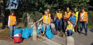Zehn Erwachsene in orangefarbenen und gelben Warnwesten stehen mit Greifzangen und mehreren vollen blauen Müllsäcken an einem Weg neben einer Mauer.