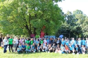 Rund achtzig Erwachsene in bunten T-Shirts und Warnwesten stehen und knien mit Eimern und Greifzangen auf einer Wiese unter einem großen Baum, dahinter Beachflags.