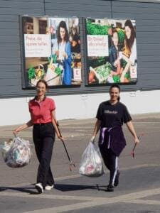 Zwei junge Frauen gehen mit Greifzangen und vollen durchsichtigen Müllsäcken über einen Parkplatz, im Hintergrund Werbeplakate eines Supermarkts.