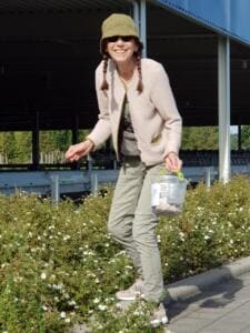 Eine lächelnde Frau mit Hut und Zöpfen sammelt Müll aus einem blühenden Beet am Rand eines Parkplatzes und hält einen Eimer.