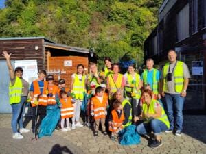 Eine Gruppe aus Kindern und Erwachsenen in gelben und orangefarbenen Warnwesten steht mit Müllsäcken und Greifzangen vor einer Holzhütte; eine Frau winkt.