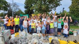 Eine große Gruppe aus Kindern und Erwachsenen reißt jubelnd die Arme hoch, davor türmen sich gefüllte Müllsäcke, Eimer und ein Anhänger.