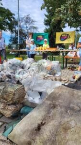 Ein großer Haufen gesammelter Abfälle aus Plastiksäcken, alten Teppichen und Planen liegt auf einem Platz, dahinter Biertische und World-Cleanup-Day-Fahnen.