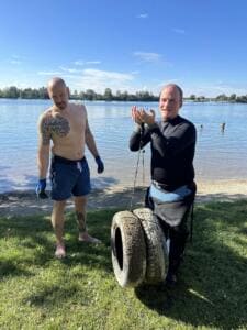 Zwei Männer stehen am Seeufer, einer im Neoprenanzug hält zwei alte Autoreifen an einem Seil, die gerade aus dem Wasser geholt wurden.