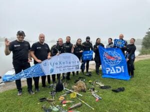 Eine Gruppe von Taucherinnen und Tauchern in Neoprenanzügen hält am Seeufer ein Banner und eine PADI-Flagge, vor ihnen liegen auf der Wiese Fundstücke aus dem Wasser.