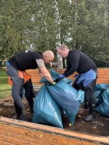Zwei Männer in Neoprenanzügen füllen auf einem Anhänger gesammelten Müll in große blaue Säcke.