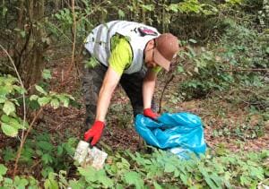 Ein Mann mit Kappe, Warnweste und roten Handschuhen zieht im dichten Unterholz ein Stück Abfall heraus und hält einen blauen Müllsack.