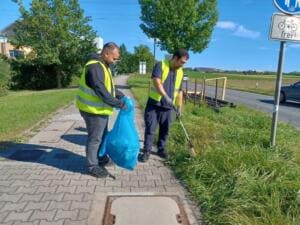 Zwei Männer in gelben Warnwesten sammeln mit Greifzange und blauem Müllsack Abfall am Rand eines Rad- und Fußwegs.