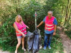 Eine Frau und ein Mann in pinkfarbenen Warnwesten stehen an einem Waldweg neben zwei vollen schwarzen Müllsäcken und einer Greifzange.