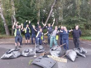 Ein gut gelauntes Team in Arbeitskleidung reckt die Arme in die Höhe, vor ihnen liegen volle schwarze Müllsäcke, ein altes Stoßstangenteil und ein Schild.