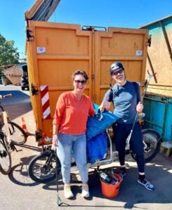 Eine Frau und ein Mann mit Fahrradhelm stehen lachend mit Greifzange und vollen Säcken vor einem orangefarbenen Container, daneben ein Lastenrad mit Eimer.
