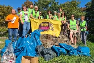 Eine Gruppe aus Erwachsenen und Kindern in gelben Warnwesten steht auf einer Steinmauer mit einer gelben World-Cleanup-Day-Fahne, davor liegen viele volle blaue Müllsäcke.
