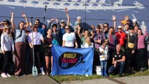 Eine gut gelaunte Gruppe winkt vor einer Wand mit Drachenboot-Motiv, vorne halten zwei Personen eine blaue World-Cleanup-Day-Fahne, einige tragen Stand-up-Paddel.