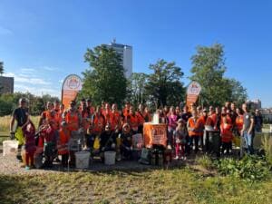 Eine große Gruppe in orangefarbenen Warnwesten steht mit Greifzangen und Eimern zwischen zwei Beachflags mit der Aufschrift Rostock Müllfrei – gemeinsam gegen Müll, dahinter ein Hochhaus.