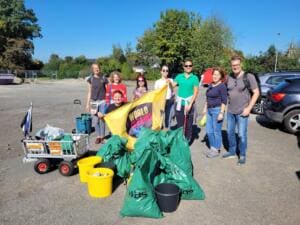 Acht Erwachsene stehen auf einem Schotterparkplatz mit einer gelben World-Cleanup-Day-Fahne, davor grüne Müllsäcke, gelbe Eimer und ein Bollerwagen.