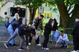 Mehrere junge Leute sammeln mit Greifzangen und Eimern Müll an einer Bushaltestelle mit dem Schild Emsterfeld, im Hintergrund ein Linienbus.