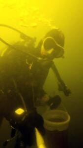 Unter Wasser im gelblich-grünen Licht hält ein Taucher mit Lampe einen Eimer für gefundenen Müll.