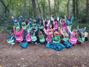 Eine Schulklasse in grünen und pinken World-Cleanup-Day-Westen kniet von hinten fotografiert im Wald und reckt die Arme und Greifzangen in die Höhe.