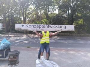 Ein Helfer in gelber Warnweste stemmt ein langes Werbeschild mit der Aufschrift Konzeptentwicklung über den Kopf, daneben Säcke und ein Bollerwagen am Straßenrand.