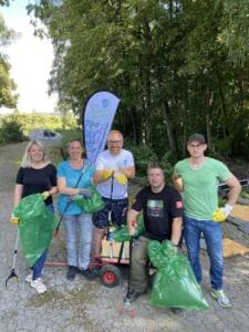 Fünf Erwachsene mit Greifzangen und grünen Müllsäcken stehen an einem Waldweg vor einer Beachflag der City Cleaners Germany, daneben ein Bollerwagen.