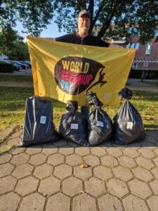 Ein Mann hält eine gelbe World-Cleanup-Day-Fahne hoch, vor ihm stehen vier prall gefüllte schwarze Müllsäcke auf einem gepflasterten Platz.