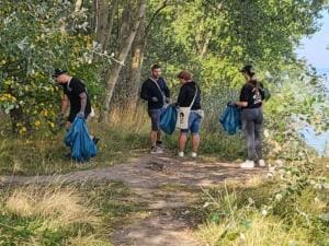 Vier Personen sammeln mit blauen Säcken Müll zwischen Büschen und Bäumen an einem Weg nahe der Ostsee.