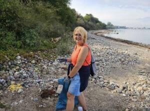 Eine lächelnde Frau mit Handschuhen und blauem Müllsack sammelt am steinigen Ostseestrand unterhalb der Steilküste.