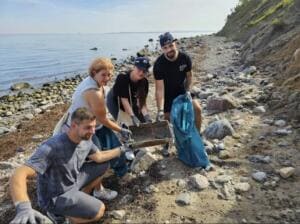 Vier Erwachsene hocken am steinigen Ostseestrand und sieben mit einem Holzsieb kleine Müllteile aus dem Sand.