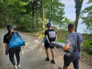 Drei junge Erwachsene gehen auf einem Waldweg an der Steilküste, eine trägt einen vollen blauen Sack, zwei Männer schleppen gemeinsam einen Betonstein.