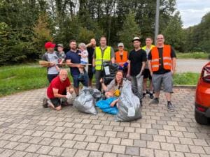 Eine Gruppe Erwachsener, einige mit kleinen Kindern auf dem Arm, steht in Warnwesten mit Besen und vollen Müllsäcken auf einem gepflasterten Parkplatz.