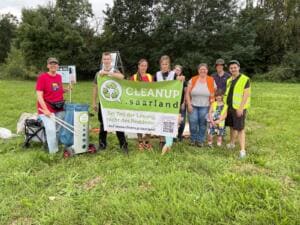 Eine Gruppe aus Erwachsenen und Kindern steht auf einer Wiese hinter einem Banner mit der Aufschrift Cleanup.saarland – Sei Teil der Lösung, nicht des Problems.