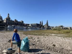 Ein Junge steht mit einem Müllsack am kiesigen Elbufer, gegenüber am anderen Ufer die Dresdner Altstadt mit Frauenkirche, Hofkirche und Ausflugsdampfern.