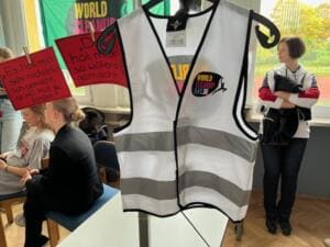 Eine weiße Warnweste mit World-Cleanup-Day-Logo hängt im Klassenzimmer, daneben Karten mit Schülerzitaten; im Hintergrund sitzen und stehen Schülerinnen.