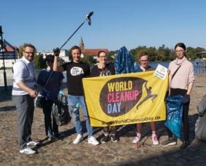 Eine Gruppe Erwachsener hält am Havelufer eine gelbe World-Cleanup-Day-Fahne und volle blaue Müllsäcke, im Hintergrund ein Kirchturm.