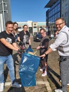 Fünf lachende Erwachsene, einer im World-Cleanup-Day-T-Shirt, halten mit Greifzangen gemeinsam einen blauen Müllsack auf einer gepflasterten Straße.