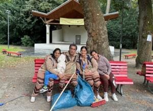 Vier Erwachsene und ein Kind sitzen lächelnd auf einer roten Parkbank vor einem Konzertpavillon, davor Müllsäcke, Besen und Greifzange.